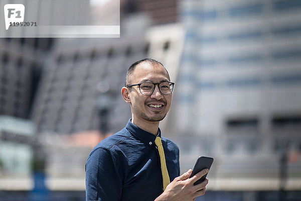 Porträt eines lächelnden Geschäftsmannes mit Smartphone vor einem Bürogebäude  New York  USA