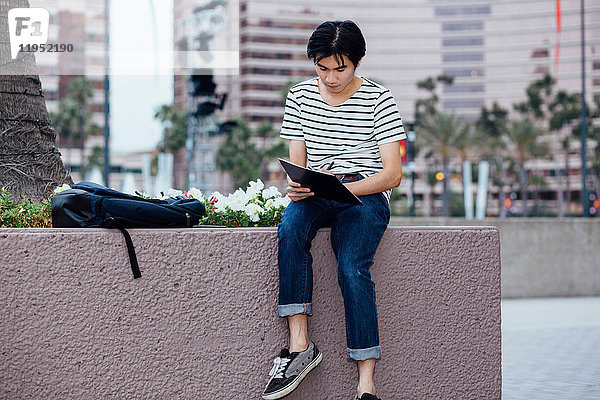Junger Mann sitzt an der Wand  im Freien  schreibt in ein Notizbuch