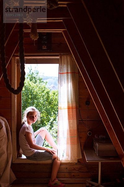 Porträt einer Teenagerin  die auf einer sonnenbeschienenen Fensterbank im Schlafzimmer sitzt