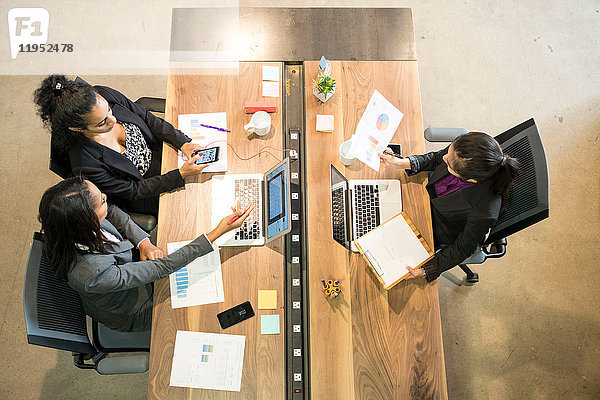 Drei Geschäftsfrauen sitzen am Schreibtisch  benutzen Laptops  halten ein Smartphone in der Hand  Blick nach oben