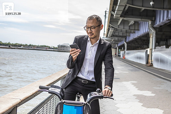 Geschäftsreisender Radfahrer betrachtet Smartphone am Wasser  New York  USA