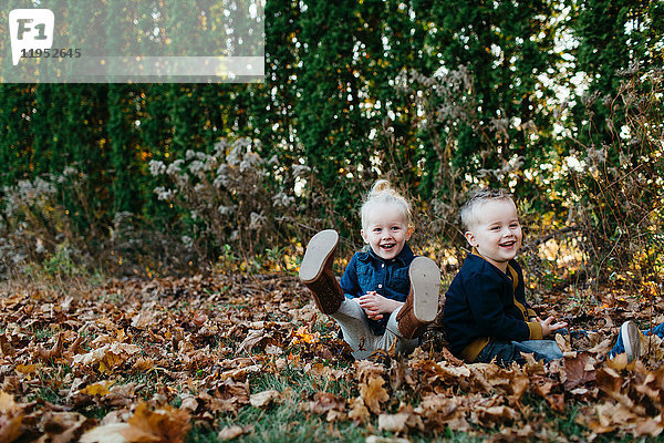 Porträt von männlichen und weiblichen Zwillingskindern  die zwischen Herbstblättern im Garten sitzen
