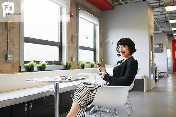 Porträt einer Geschäftsfrau  die in einer Büroumgebung am Tisch sitzt  ein Smartphone in der Hand hält und lächelt