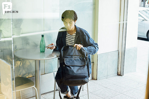 Junger Mann sitzt an Café-Tisch  sortiert Tasche durch  hält Smartphone