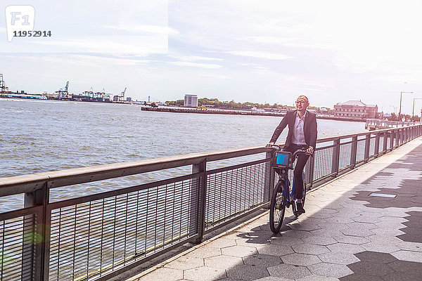 Mittelgroßer erwachsener Geschäftsmann mit dem Fahrrad entlang der Uferpromenade des Stadtflusses