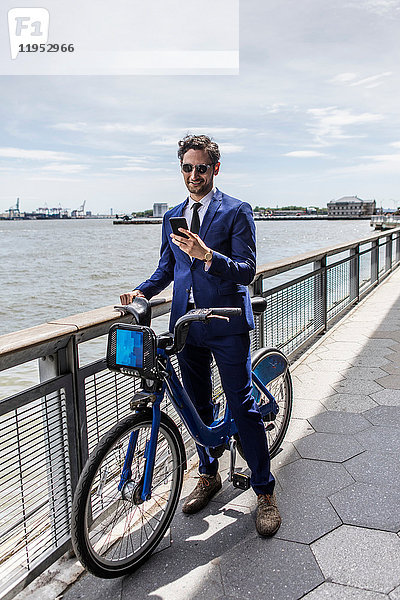Junger Geschäftsmann auf dem Fahrrad schaut auf Smartphone am Ufer des Stadtflusses