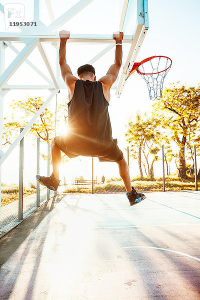 Junger Mann auf dem Basketballplatz  auf Basketballnetzrahmen schwingend  Rückansicht