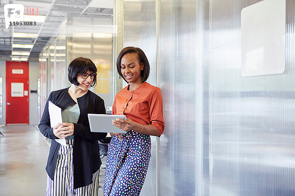 Zwei Geschäftsfrauen stehen im Bürokorridor  schauen auf das digitale Tablet und lächeln