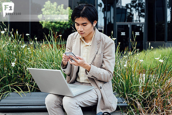 Junger Mann sitzt im Freien  benutzt Laptop  hält Smartphone