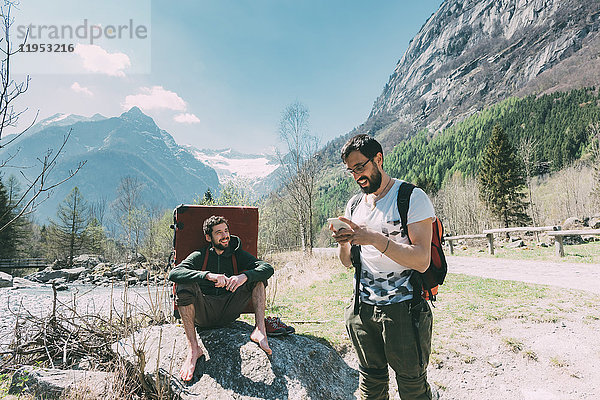 Zwei männliche Freunde mit Bouldermatten-Rucksack beim Blick auf Smartphone am Flussufer  Lombardei  Italien