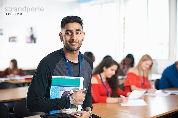 Porträt eines Schülers im Klassenzimmer  der ein Lehrbuch in der Hand hält und lächelnd in die Kamera schaut