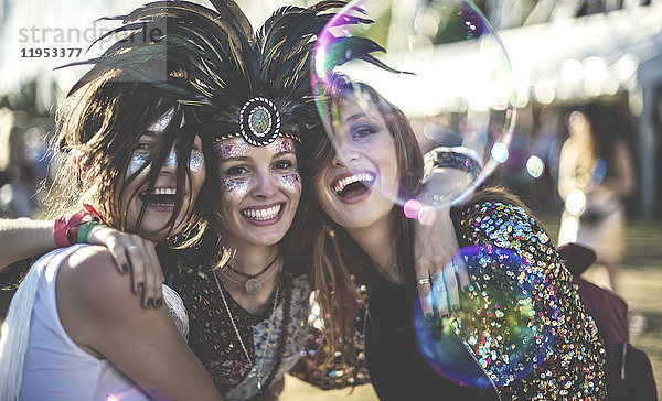Drei junge Frauen bei einem Sommer-Musikfestival mit Federkopfschmuck und bemalten Gesichtern  die in die Kamera lächeln.