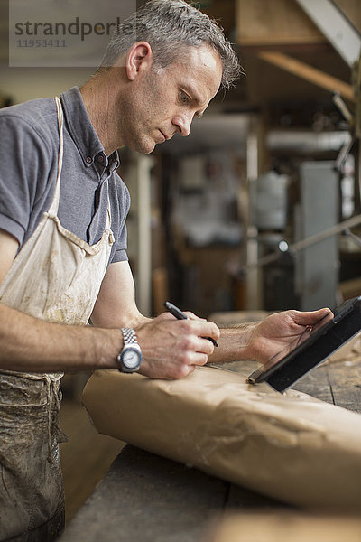 Ein Mann in einer Möbelrestaurierungswerkstatt  der ein digitales Tablett benutzt und auf ein mit braunem Verpackungspapier umhülltes Päckchen schreibt