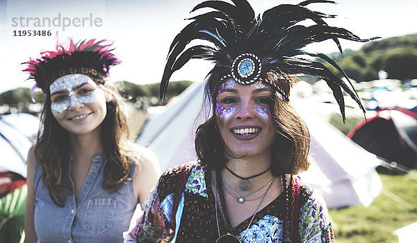 Zwei lächelnde junge Frauen bei einem Sommer-Musikfestival mit bemaltem Gesicht und Federkopfschmuck zwischen Zelten stehend.