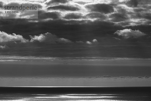 Blick vom Land über den Ozean  bei schwachem Licht bis zum Horizont. Sonnenlichtflecken auf der Wasseroberfläche und dunkle Gewitterwolken über uns.