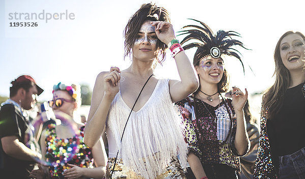 Zwei lächelnde junge Frauen bei einem Sommer-Musikfestival mit bemaltem Gesicht  mit Federkopfschmuck  stehen in der Menge und schauen in die Kamera.