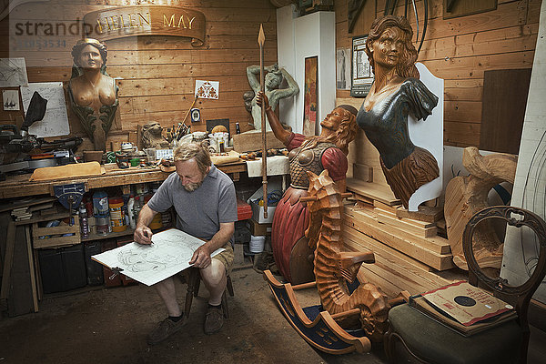 Ein Handwerker  Holzarbeiter sitzt auf einem Hocker in einer Werkstatt und arbeitet an einer Zeichnung  die er mit Holzkohle skizziert. Umgeben von geschnitzten und bemalten weiblichen Schiffsgestalten aus Holz.