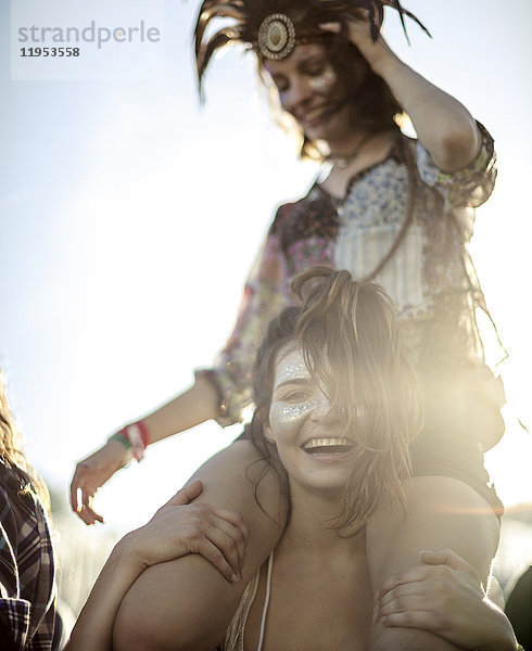 Junge Frau bei einem Sommer-Musikfestival  die eine Freundin auf den Schultern trägt  mit geschminktem Gesicht und einem Lächeln in die Kamera.