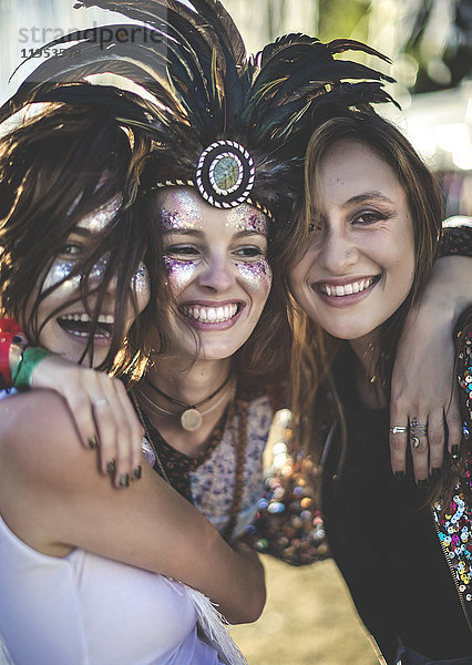 Drei junge Frauen bei einem Sommer-Musikfestival mit Federkopfschmuck und bemalten Gesichtern  die in die Kamera lächeln.
