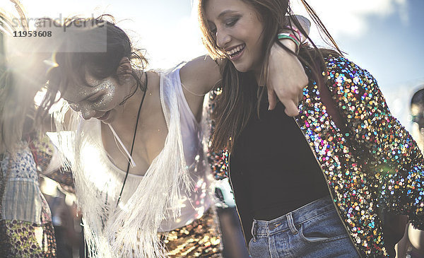 Zwei junge Frauen bei einem Sommer-Musikfestival  die Pailletten tragen  mit gemalten Gesichtern  die lachen und tanzen.
