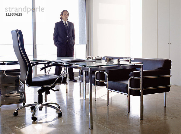 Geschäftsmann in dunklem Anzug  der drinnen in einem Büro hinter einem Schreibtisch mit Stühlen steht.
