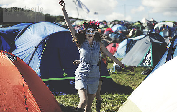 Junge Frau bei einem Sommer-Musikfestival mit geschminktem Gesicht  Federkopfschmuck  steht in der Nähe des Campingplatzes  umgeben von Zelten  den Arm erhoben  lächelnd.