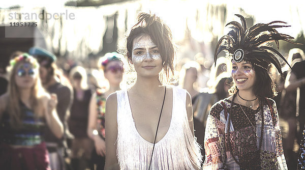 Zwei lächelnde junge Frauen bei einem Sommer-Musikfestival mit bemaltem Gesicht  mit Federkopfschmuck  stehen in der Menge und schauen in die Kamera.