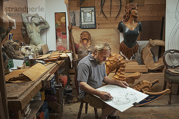 Ein Handwerker  Holzarbeiter sitzt auf einem Hocker in einer Werkstatt und arbeitet an einer Zeichnung  die er mit Holzkohle skizziert. Umgeben von geschnitzten und bemalten weiblichen Schiffsgestalten aus Holz.