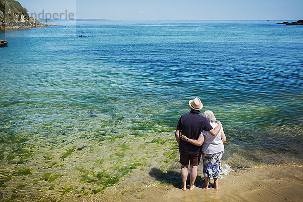 Rückansicht eines älteren Ehepaares  das mit den Armen umeinander steht und auf das leuchtend blaue und türkisfarbene Wasser  den Ozean  blickt.