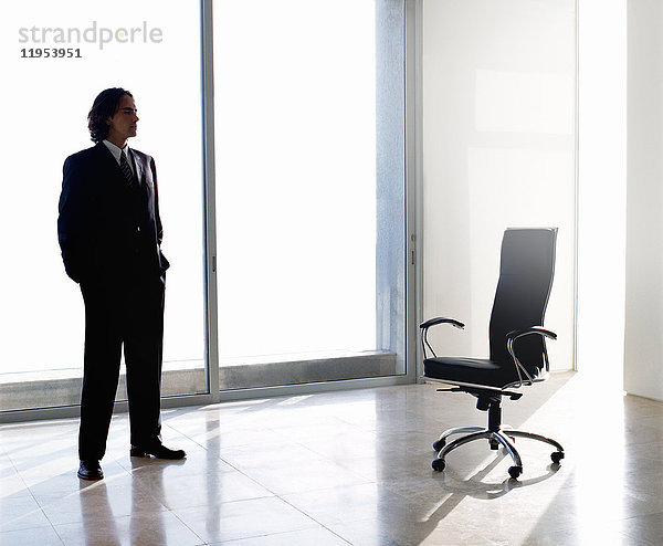 Geschäftsmann in dunklem Anzug steht drinnen neben dem Bürostuhl vor dem Fenster  die Hände in den Taschen.