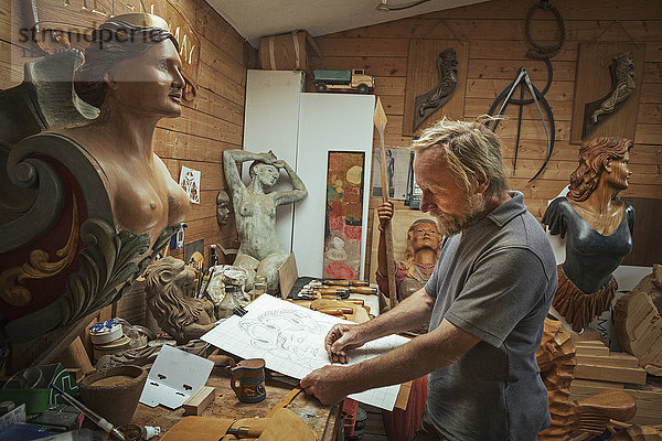 Ein Handwerker  Holzarbeiter  der in einer Werkstatt an einer Bank steht und an einer Zeichnung arbeitet  die er mit Holzkohle skizziert. Umgeben von geschnitzten weiblichen Schiffsgestalten aus Holz.