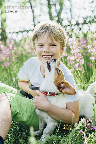 Porträt eines lächelnden kleinen Jungen  der mit seinem Hund auf einer Wiese im Garten sitzt.