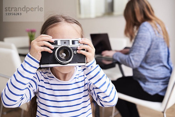 Kleines Mädchen fotografiert mit der Kamera  während ihre Mutter im Hintergrund am Laptop arbeitet.