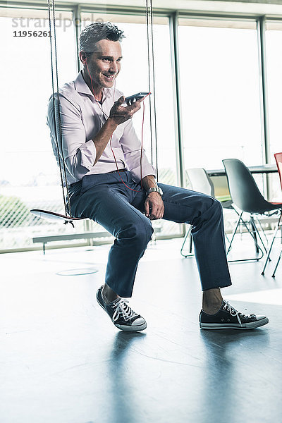 Geschäftsmann im Büro sitzend auf Schaukel  mit Smartphone