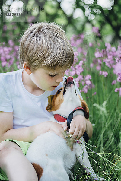 Kleiner Junge sitzt mit seinem Hund auf einer Wiese im Garten.