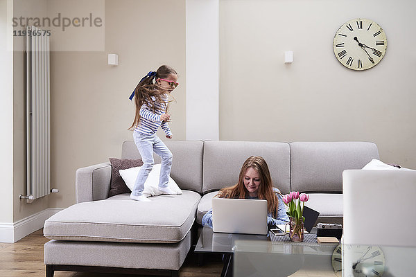 Kleines Mädchen mit Sonnenbrille  das auf dem Sofa springt  während ihre Mutter am Laptop am Couchtisch arbeitet