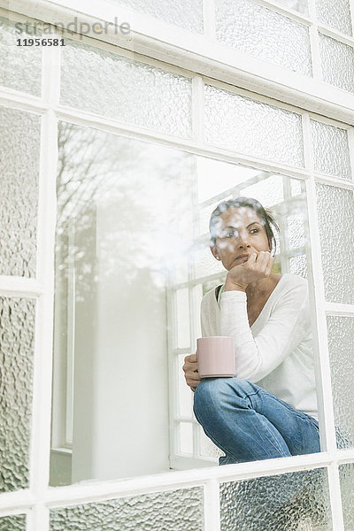 Nachdenkliche Frau mit einer Tasse Kaffee  die aus dem Fenster schaut.