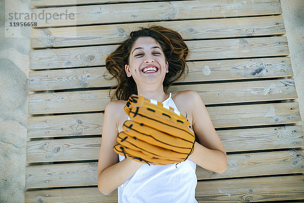 Junge Frau auf Holzweg liegend  lachend mit Baseballhandschuhen