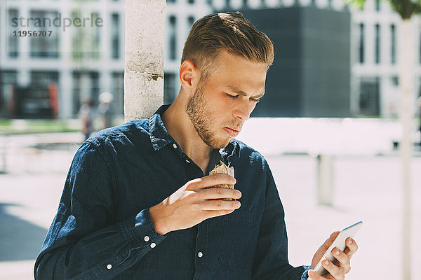 Ein junger Geschäftsmann beim Mittagessen draußen  während er sein Handy ansieht.