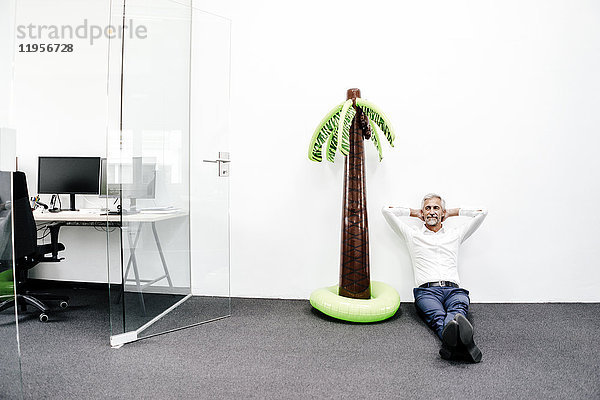 Lächelnder reifer Geschäftsmann sitzt neben aufblasbarer Palme im Büro