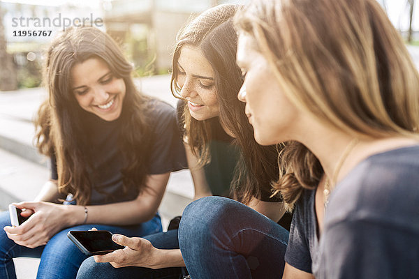 Drei glückliche junge Frauen sitzen draußen und schauen auf ihr Handy.