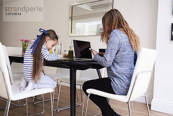 Kleines Mädchen beim Zeichnen am Tisch  während ihre Mutter mit dem Handy telefoniert.