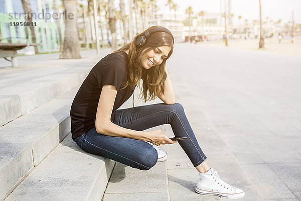 Lächelnde junge Frau sitzt auf einer Treppe und hört Musik.
