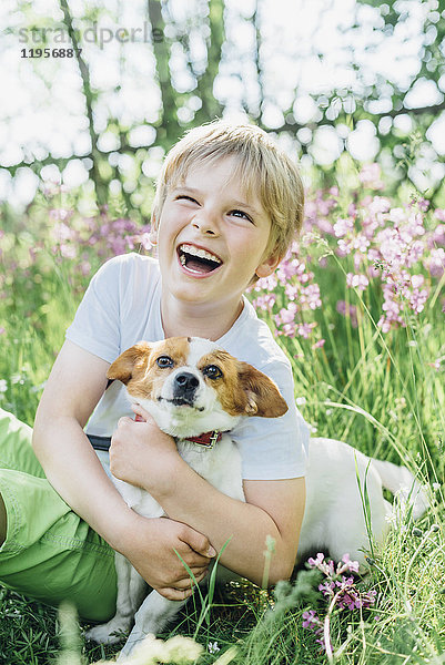 Porträt eines lachenden kleinen Jungen  der mit seinem Hund auf einer Wiese im Garten sitzt.