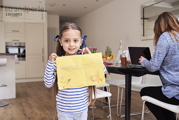 Porträt eines lächelnden Mädchens mit Zeichnung  während ihre Mutter im Hintergrund am Laptop arbeitet.