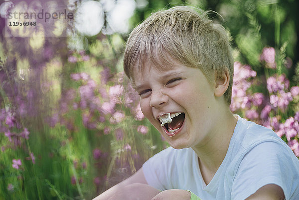 Porträt eines lachenden kleinen Jungen mit Kaugummi auf einer Wiese im Garten sitzend