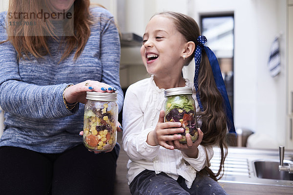 Lachendes kleines Mädchen mit einem Einmachglas mit gemischtem Salat neben ihrer Mutter.