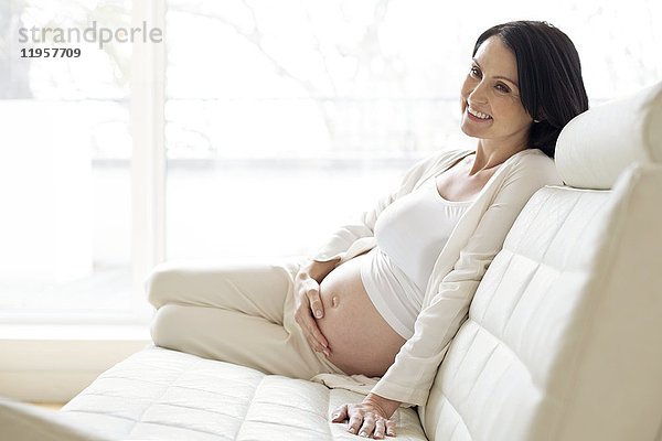 MODELL FREIGEGEBEN. Schwangere Frau sitzt auf dem Sofa und berührt ihren Bauch.