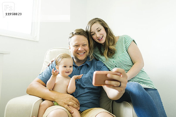 Mutter und Vater mit einem kleinen Mädchen (12-17 Monate) sitzen im Wohnzimmer und machen ein Selfie