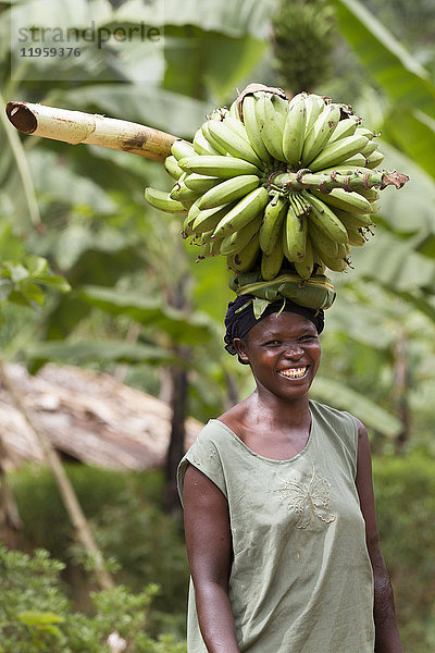 Porträt einer lächelnden Frau  die ein großes Bündel Bananen auf dem Kopf trägt  Uganda  Afrika